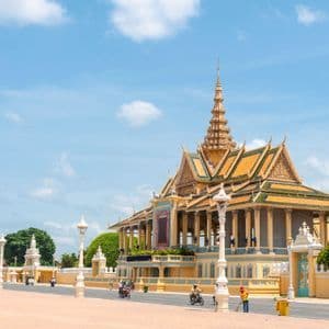 An ornate yellow palace with a multi-tiered golden roof stands beside a paved square under a bright blue sky.