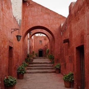 Uno stretto vicolo in pietra con mura rosso terracotta e archi conduce a una scalinata fiancheggiata da piante in vaso.