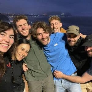 Eine WeRoad-Gruppenreise mit lachenden Freunden, die nachts ein Gruppen-Selfie aufnehmen, mit Blick auf eine beleuchtete Küste.