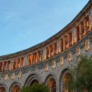Un grande edificio in pietra ricurvo con colonne e archi classici, illuminato da luci calde contro un cielo crepuscolare.