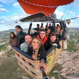 Un gruppo WeRoad si scatta un selfie all'interno di una cesta di mongolfiera che sorvola una valle rocciosa con altre mongolfiere nel cielo.