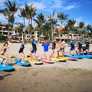 A WeRoad group trip taking a surfing lesson on a sandy beach, practicing on colorful surfboards under palm trees.