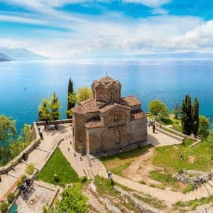 A panoramic view of an old stone church on a cliffside overlooking a vast blue lake, with distant mountains under a cloudy sky.