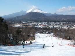 Persone che sciano e fanno snowboard su un'ampia pista con una città e un grande vulcano innevato sullo sfondo.