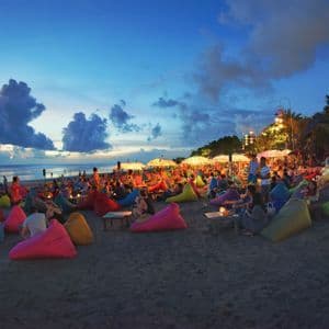 Un groupe WeRoad se détend sur des poufs colorés sur une plage de sable au crépuscule, avec l'océan en arrière-plan.