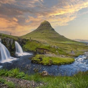 Ein mehrstufiger Wasserfall stürzt in einen Fluss mit einem markanten kegelförmigen grünen Berg im Hintergrund unter einem Sonnenuntergangshimmel.