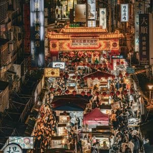 Eine Vogelperspektive auf einen belebten asiatischen Nachtmarkt, auf dem Menschen zwischen Ständen unter beleuchteten Schildern und roten Laternen umhergehen.