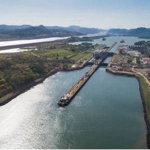 Una vista aérea de las esclusas del Canal de Panamá, con un remolcador entrando al canal y buques de carga a lo lejos.