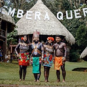 Vier Embera Quera Menschen in traditioneller Kleidung und Körperbemalung stehen für ein Foto vor strohgedeckten Hütten.