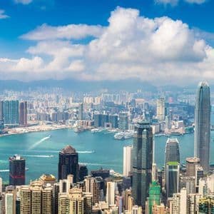 Una vista panoramica su uno skyline denso di città costiera, con moderni grattacieli che circondano un ampio porto sotto un cielo azzurro brillante con nuvole.