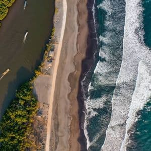 Eine Luftaufnahme eines Flusses mit Booten neben einem Sandstrand, der an den türkisfarbenen Ozean mit weißen Wellen grenzt.