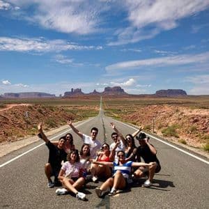 Un gruppo in viaggio con WeRoad posa al centro di una strada desertica che si estende verso lontane mesa sotto un cielo blu.