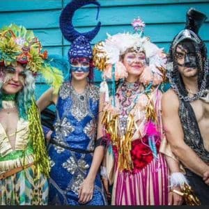 A WeRoad group trip of four people posing in elaborate, colorful costumes with headdresses and face paint against a blue wall.