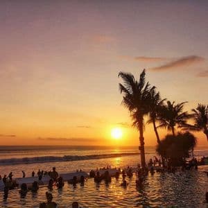 Voyage en groupe WeRoad : des personnes dans une piscine à débordement, admirant le coucher de soleil sur l'océan avec des palmiers en silhouette.