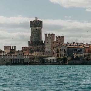 A large stone castle with towers and battlements stands on the shore of a choppy lake under a cloudy sky.