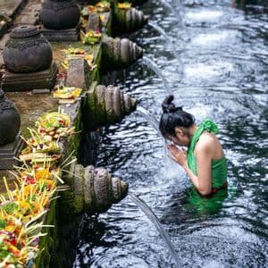 A woman in a green top performs a purification ritual, bowing her head under a stream of water from a stone fountain.