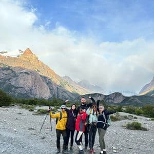Un viaggio di gruppo WeRoad con attrezzatura da trekking posa in una valle montuosa rocciosa con un debole arcobaleno sullo sfondo.