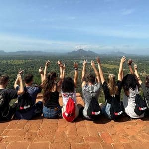 Eine WeRoad-Gruppe steht mit dem Rücken zur Kamera und erhobenen Armen auf einem hohen Aussichtspunkt, mit Blick auf ein weites grünes Tal und Berge.