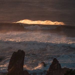 Una grande onda oceanica si infrange nell'acqua, la sua cresta che risplende di luce solare dorata sopra le rocce scure in primo piano.