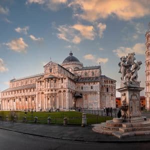 Una vista panoramica di una grande cattedrale, una torre pendente e un battistero accanto a un prato verde sotto un cielo parzialmente nuvoloso.