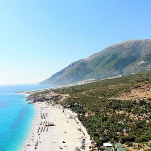 Vue aérienne d'une longue plage de sable et d'eaux turquoise, bordée par une grande montagne verte sous un ciel bleu azur.
