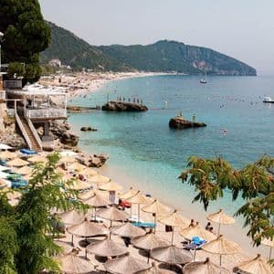 Vue aérienne d'une plage bondée, avec de nombreux parasols en paille, une eau turquoise et limpide, et un littoral montagneux boisé.