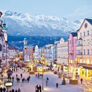 Una vivace via pedonale affollata, fiancheggiata da edifici colorati e alberi illuminati, con uno sfondo di montagne innevate.