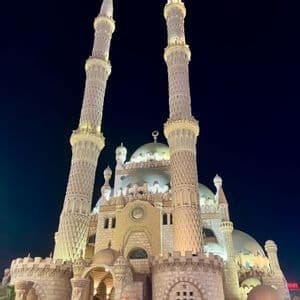 Una moschea in pietra ornata con grandi cupole e due alti minareti è illuminata da luci contro un cielo notturno scuro.