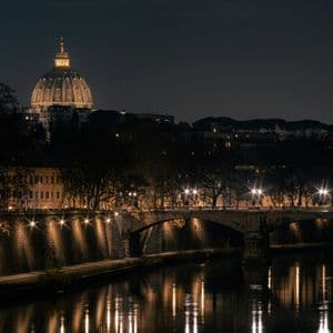 Eine beleuchtete Kuppel erhebt sich nachts über eine Stadtsilhouette, während sich eine Steinbrücke und Straßenlaternen im darunterliegenden Fluss spiegeln.