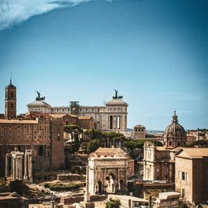A high-angle view of ancient ruins, including arches and columns, with a historic city skyline under a blue sky.
