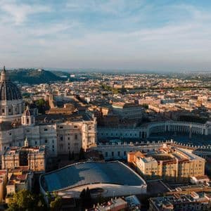 An aerial view of a grand basilica with a large dome and a vast colonnaded piazza, overlooking a sprawling city at sunrise.