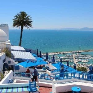 Edifici bianchi con terrazze e ombrelloni blu si affacciano su un porto turistico pieno di barche accanto al mare blu.