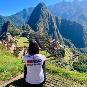 Un capogruppo WeRoad è seduto su una collina erbosa, con vista su un'antica città di rovine di pietra incastonata tra alte montagne verdi.
