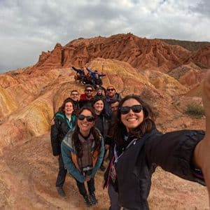 A smiling woman takes a selfie with her WeRoad group trip among colorful red and yellow rock formations under a cloudy sky.