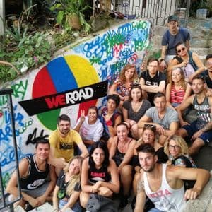 A WeRoad group trip poses for a photo on outdoor concrete stairs next to a wall with colorful graffiti and a WeRoad logo.