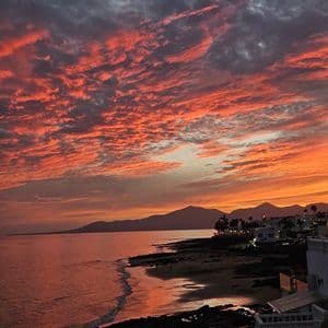 Nubes dispersas rojas y naranjas durante el atardecer sobre el océano, con una costa y montañas silueteadas al fondo.