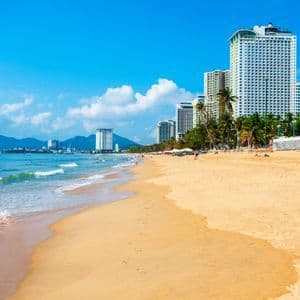 Ein breiter Sandstrand mit sanften Meereswellen, dahinter eine moderne Stadtskyline mit Hochhäusern und Palmen unter blauem Himmel.