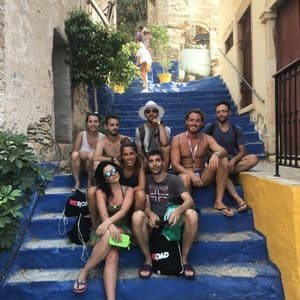 Eine WeRoad-Reisegruppe von acht Personen sitzt gemeinsam auf einer breiten, blau gestrichenen Treppe in einer engen Dorfgasse.