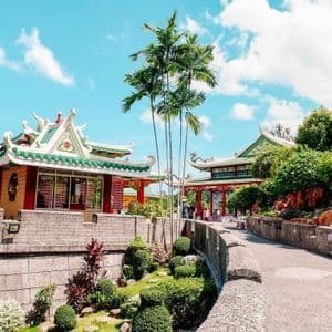Templi sontuosi con tetti di tegole verdi circondati da giardini lussureggianti, palme e sentieri in pietra sotto un cielo blu.