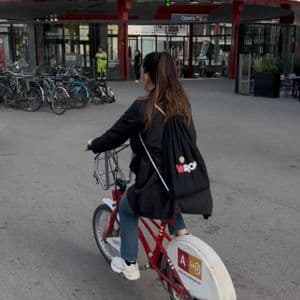 Una mujer vista de espaldas, con una mochila negra de WeRoad, monta en una bicicleta roja por una plaza adoquinada de la ciudad.
