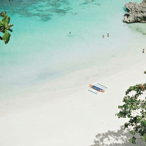 Veduta aerea di una spiaggia di sabbia bianca con persone che nuotano in acque cristalline turchesi e una canoa a bilanciere sulla riva.