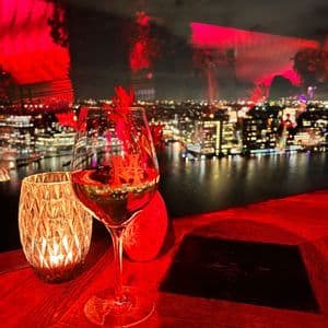 Una copa de vino y una vela en una mesa con luz roja, con vistas a un horizonte urbano iluminado al otro lado de un río por la noche.