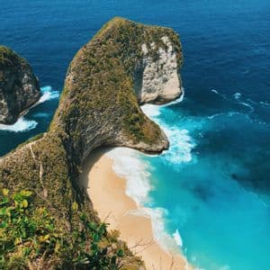 Una vista aérea de una gran formación de acantilado con cima verde que se adentra en el océano turquesa y azul, encerrando una playa de arena debajo.