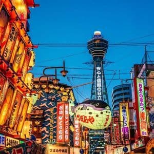 Letreros y faroles iluminados, incluido un gran pez globo, bordean una calle de la ciudad con una torre visible al fondo contra un cielo azul crepuscular.