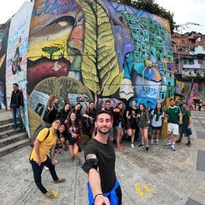 A WeRoad group trip poses for a selfie in front of a large, colorful graffiti mural in a hillside neighborhood.