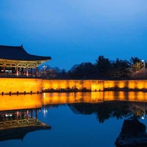 Un tradizionale padiglione e muro dell'Asia orientale, illuminato da luci gialle, che si riflette su un laghetto calmo al crepuscolo.