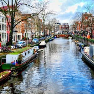 Boote liegen an einem Stadtkanal, der von farbenfrohen Gebäuden gesäumt wird, während Menschen auf der angrenzenden Straße unter einem teils bewölkten Himmel spazieren.