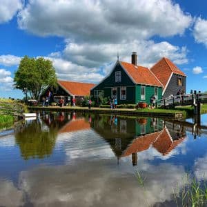 Casas tradicionales verdes y oscuras con tejados rojos, reflejándose en un canal tranquilo bajo un cielo azul con nubes blancas.