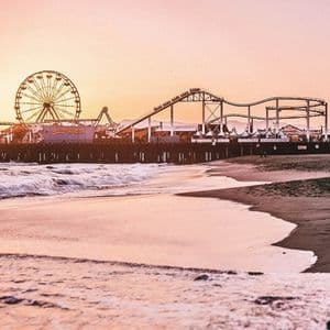 Ein Freizeitpark mit Riesenrad und Achterbahn auf einem Pier, von einem Sandstrand bei Sonnenuntergang aus gesehen.