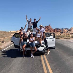 Eine WeRoad-Gruppenreise posiert für ein Foto auf und um einen weißen SUV auf einer Straße in einer Wüste mit roten Felsformationen.